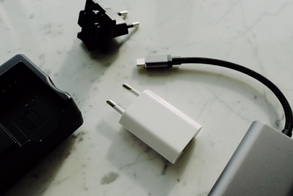 A close-up image showcasing electronic accessories placed on a white marble surface, including a black power adapter with a detachable plug, a white USB wall charger, a black power cord with a connector, and a silver-colored laptop partially visible in the corner. The setting appears to be an indoor workspace with natural lighting reflecting subtly on the marble, emphasizing the textures of the plastic and metal components. The focus is on the neatly arranged electronic devices and accessories, with no outdoor elements or gardening tools present, indicating a context unrelated to gardening, landscaping, or outdoor maintenance.