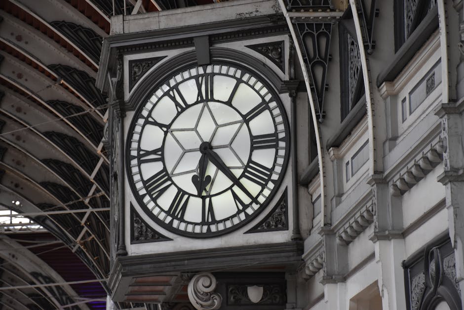 A large, ornate clock face with Roman numerals and black clock hands mounted on the exterior of a historic building. The clock's white face is framed by decorative black metalwork, and behind it, a portion of the building's decorative architecture with intricate moldings and elegant stone details is visible. Above the clock, a metal canopy with a curved, ribbed design extends outward, supported by ornate brackets. The scene is well-lit with natural daylight, highlighting the textures of the clock’s face, the detailed architectural elements, and the metallic structure above, creating a classic cityscape ambiance typical of a historic station or public building.